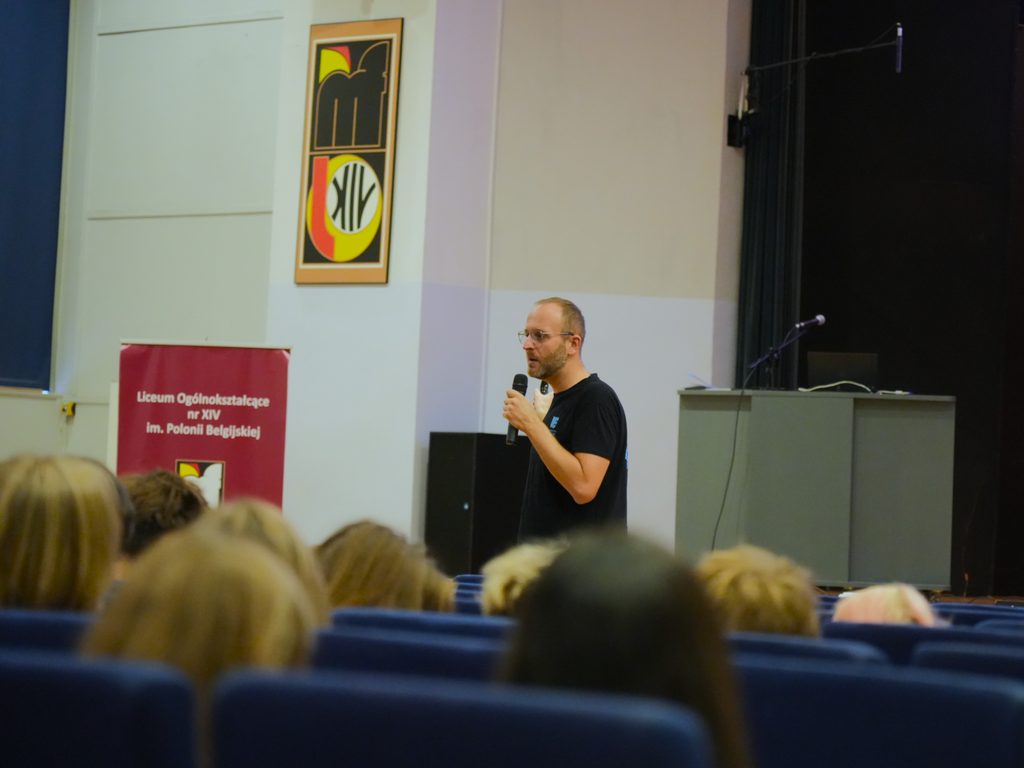 Aula XIV Liceum Ogólnokształcącego we Wrocławiu. Krystian Piątek z Sekcji Promocji Kształcenia Uniwersytetu Ekonomicznego we Wrocławiu, prezentuje ofertę edukacyjną uczelni. W sali przed prelegentem znajduje się około 50 uczniów klas maturalnych. W tle widać logotyp XIV Liceum Ogólnokształcącego we Wrocławiu. 
